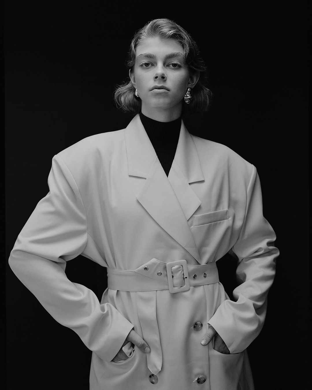 Monochrome portrait of a young adult woman in a light-colored, oversized blazer with hands in pockets against a dark background.