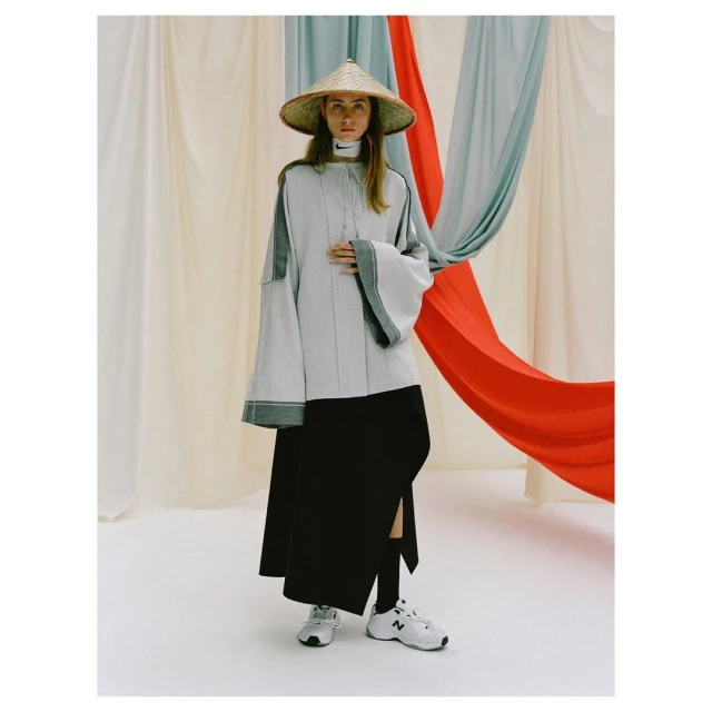 Adult female fashion model poses in a light-grey top, straw hat, and black skirt with a slit, standing against draped fabric.