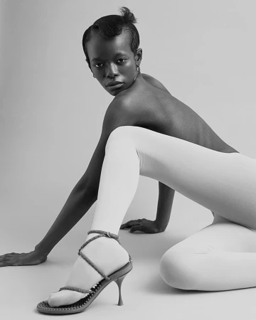 Black woman kneeling on a light backdrop wearing white tights and strapped high heels with her back partially exposed