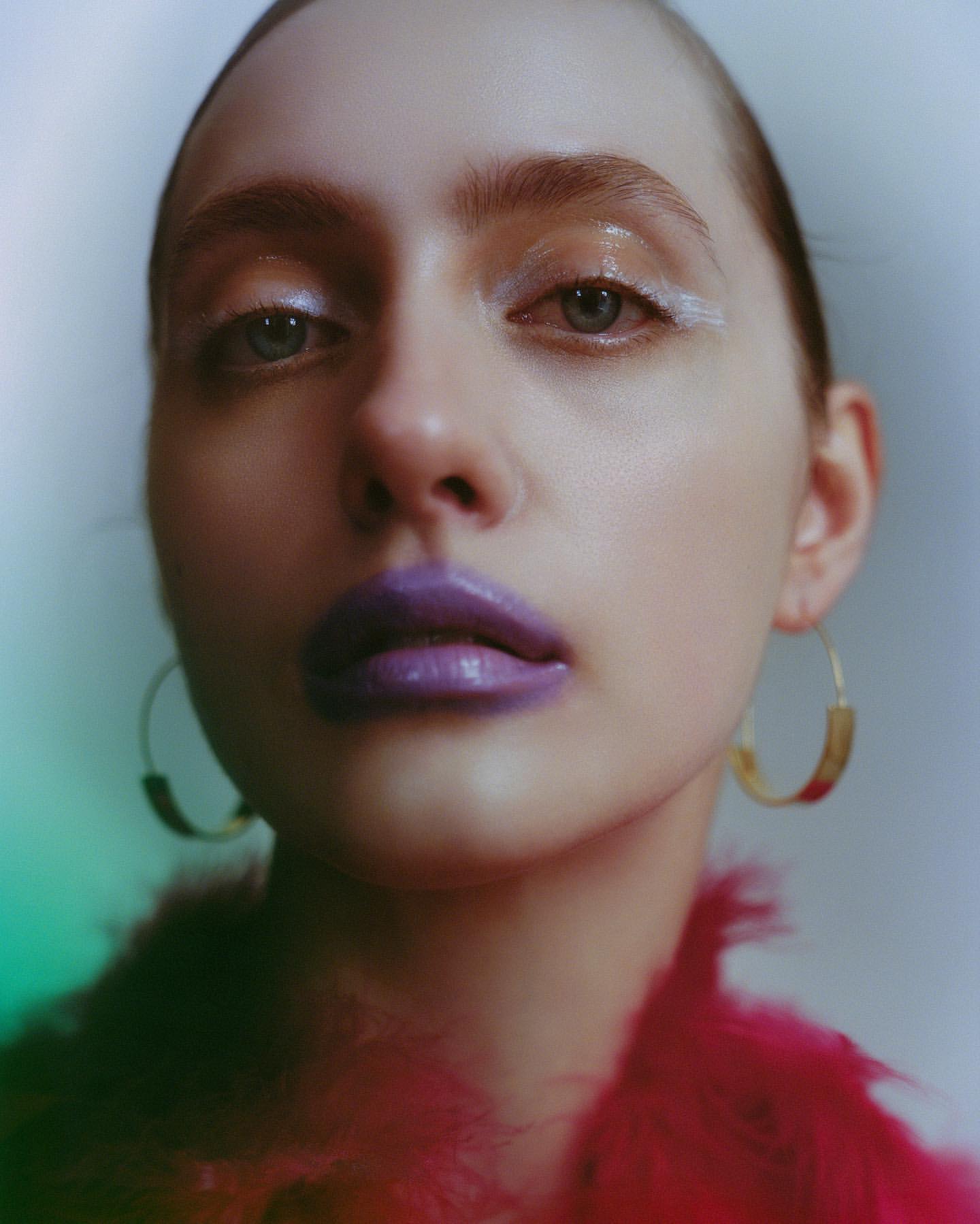 Close-up portrait of a woman with pale skin, bold purple lipstick, and shimmering eyeshadow against a blurred, multicolored background.
