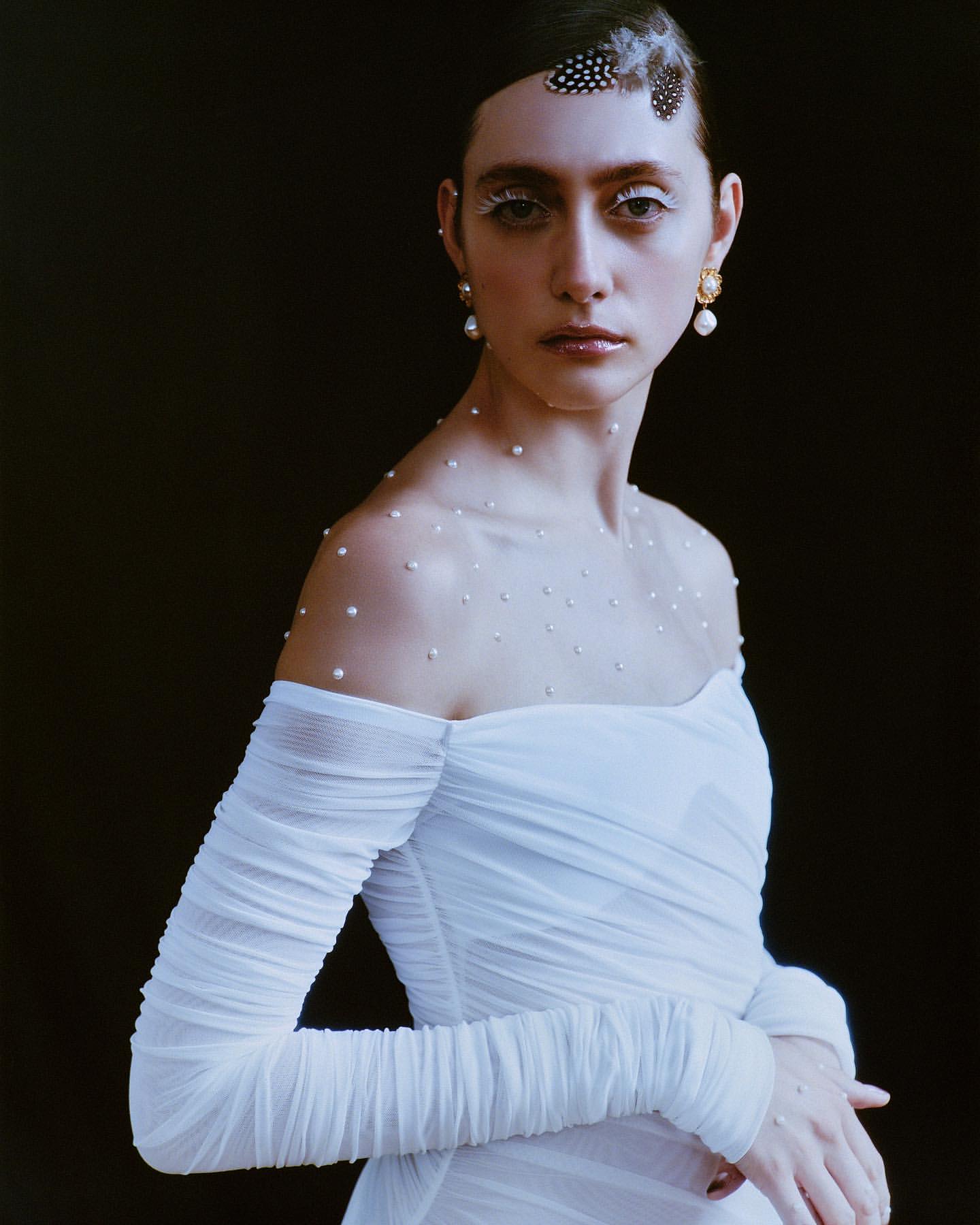 Elegant woman in white gown with pearl accents, striking pose against a dark backdrop, creating a sophisticated fashion portrait.