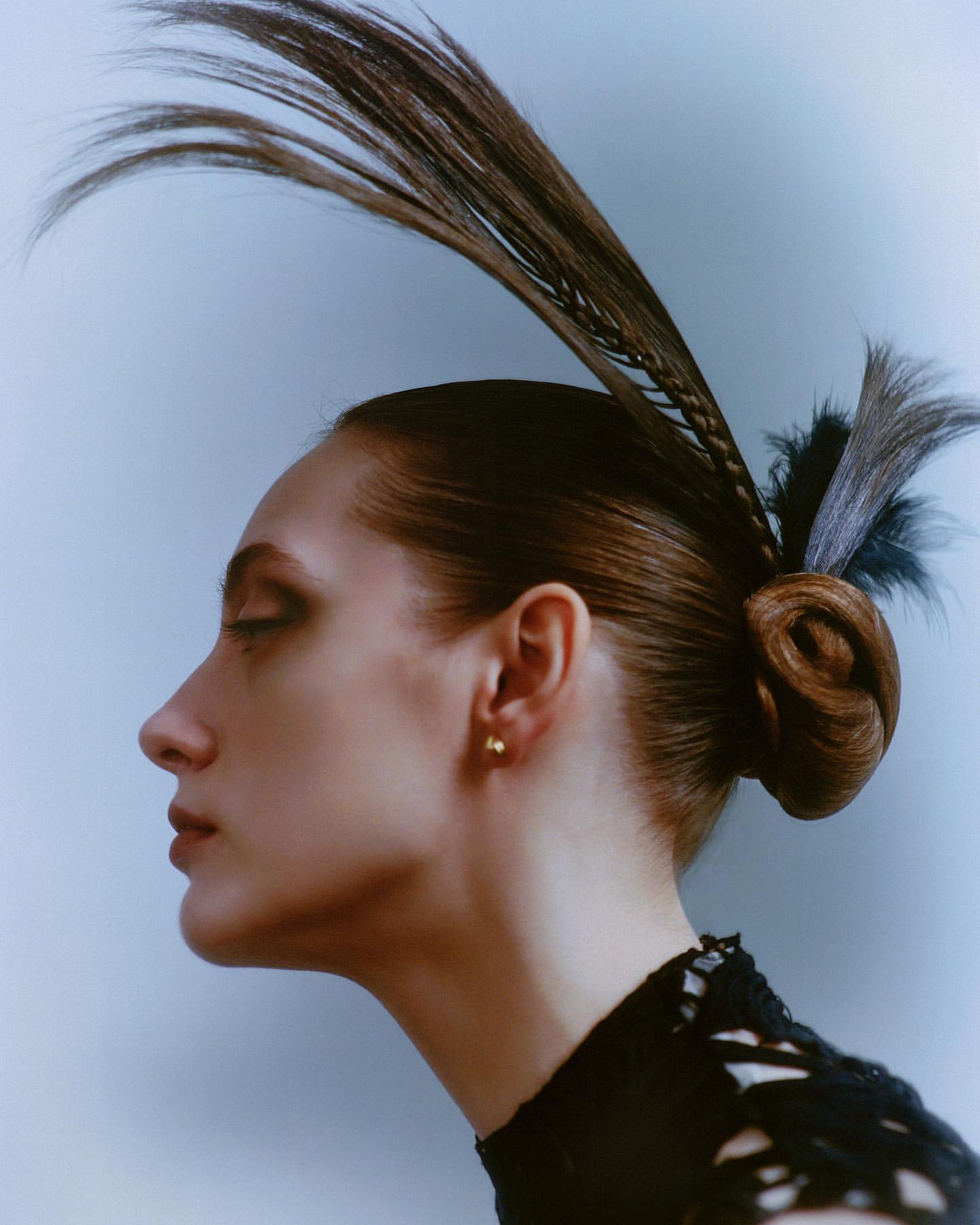 Profile of a woman with intricate brown updo, including braids and feather-like protrusions, and a black lace top against light background.
