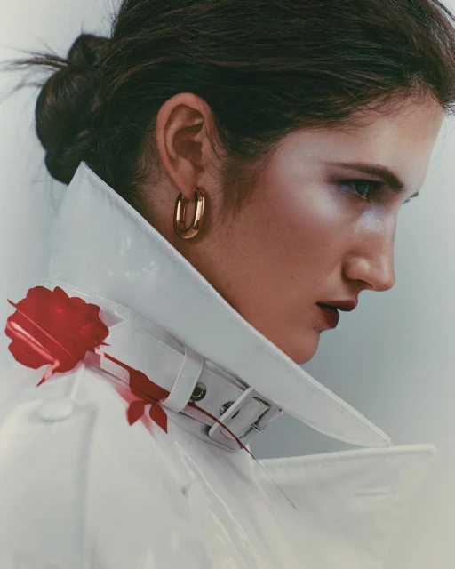 Close-up studio portrait of a young adult female model wearing a white vinyl coat with a red rose, posing against a grey background.