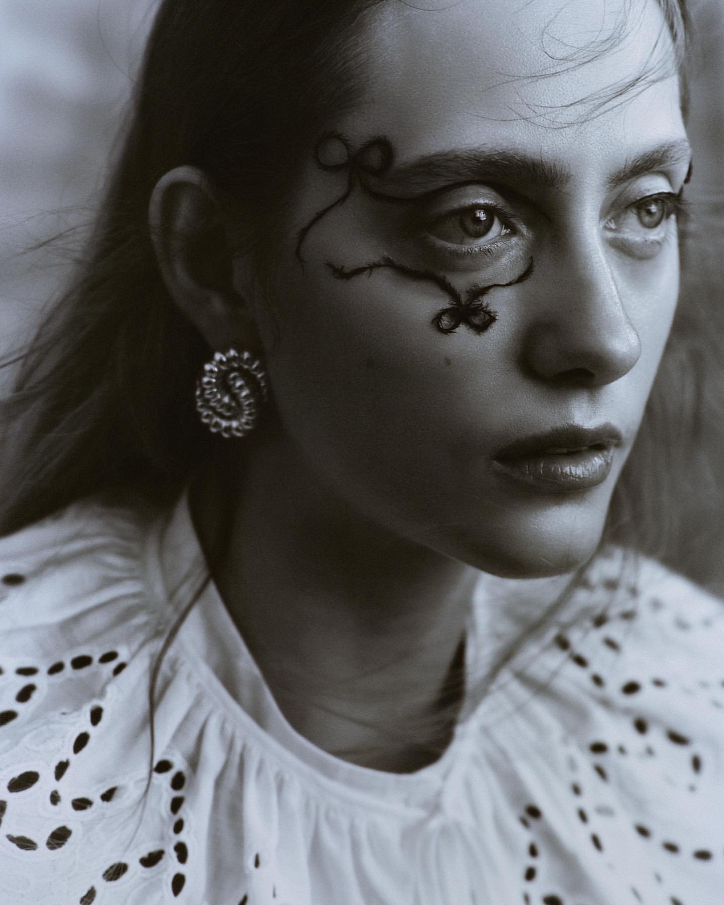 Close-up black and white portrait of a model with decorative makeup, statement earrings, and a white eyelet top in a studio setting.