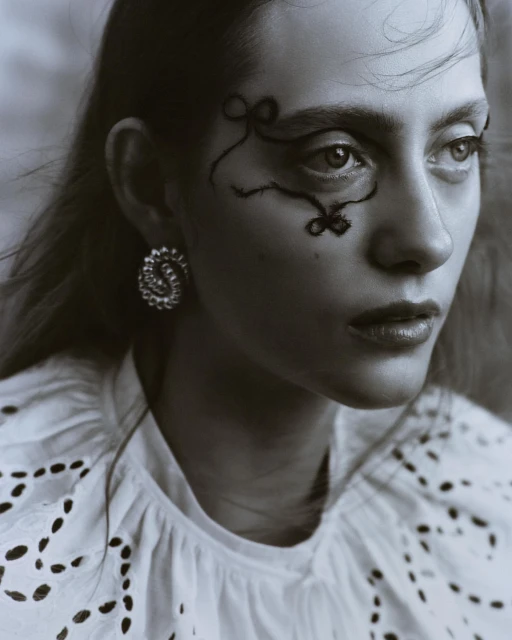 Close-up black and white portrait of a model with decorative makeup, statement earrings, and a white eyelet top in a studio setting.