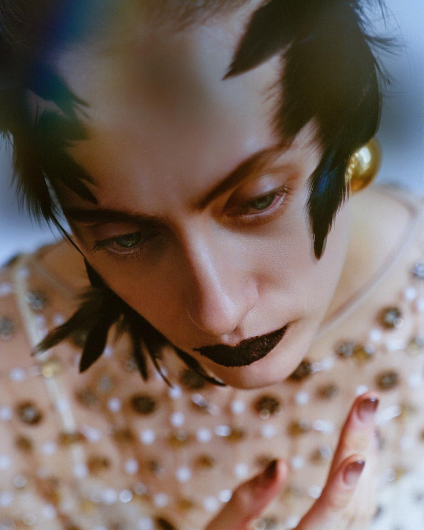 Portrait of a woman with dark lipstick and feathers in her hair, wearing an embellished top, looking down with a serious expression.