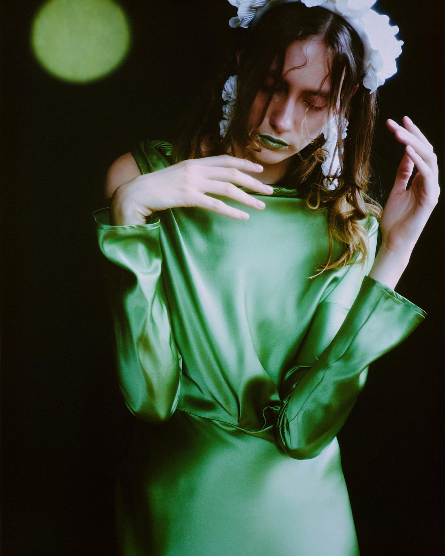 A young woman with light skin in a green dress poses in front of a dark backdrop with a round spotlight. She wears a white floral headpiece.