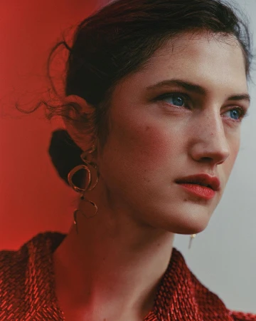 Portrait of a woman with blue eyes wearing ornate gold earrings and a red textured top against a split red and white background