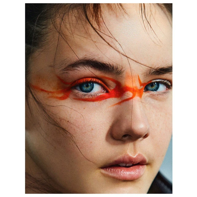 Close-up portrait of a woman with bright blue eyes and a bold orange makeup design around her eyes, creating a striking and artistic look.
