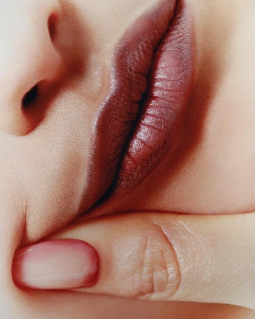 Close-up of a woman's face featuring dark red lipstick and a manicured fingernail touching her lip; beauty, fashion, or skincare.