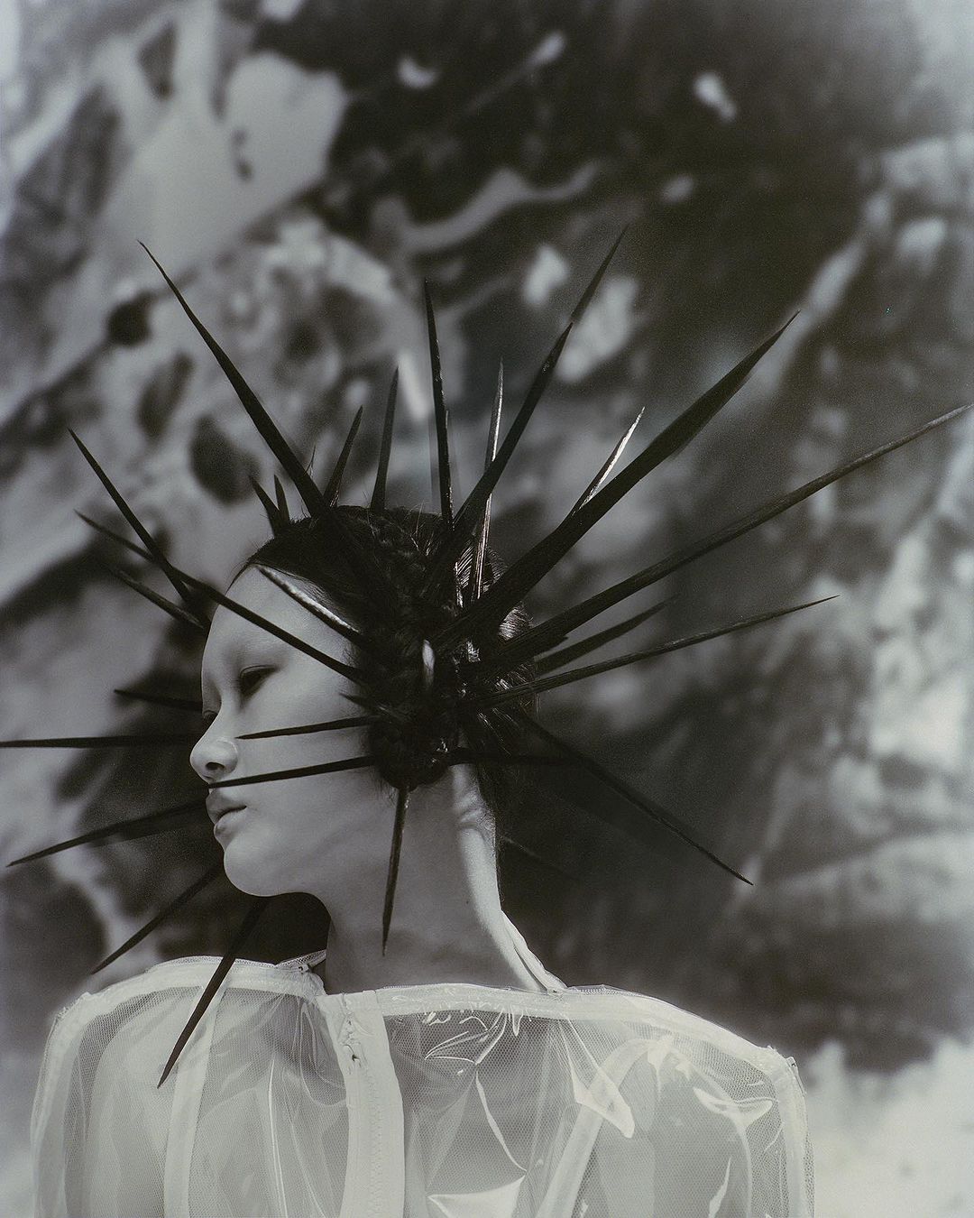 Striking black and white portrait of a woman with an elaborate spiky headpiece, wearing a translucent top against a blurred background.