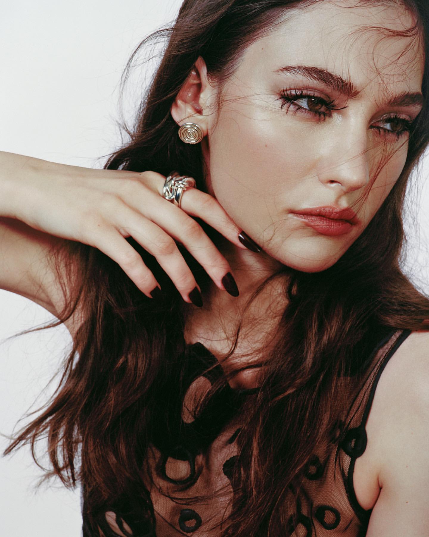 Woman with long brown hair poses in a sheer black top, wearing silver earrings and a silver ring, in a close-up studio shot.