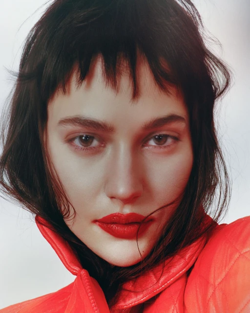Close-up studio portrait of a female fashion model with red lipstick and short dark hair wearing a red quilted jacket.