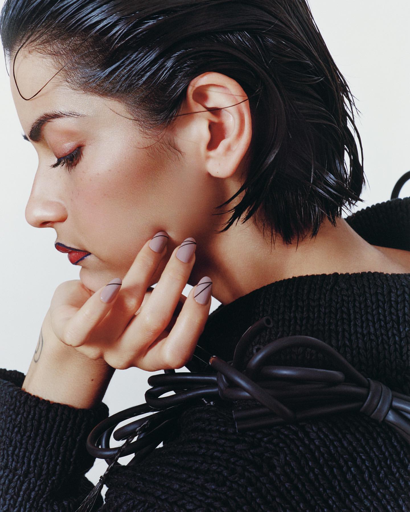 Portrait of a woman with wet dark hair and creative makeup wearing a knitted sweater holding a black electrical wire bundle indoors.