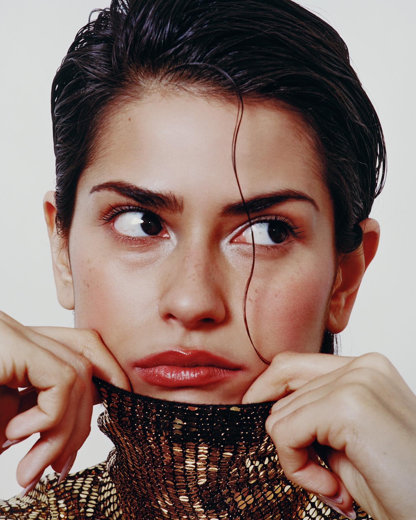 Close-up portrait of a young woman with dark hair slicked back, wearing a shimmering gold turtleneck and tilting her chin up.