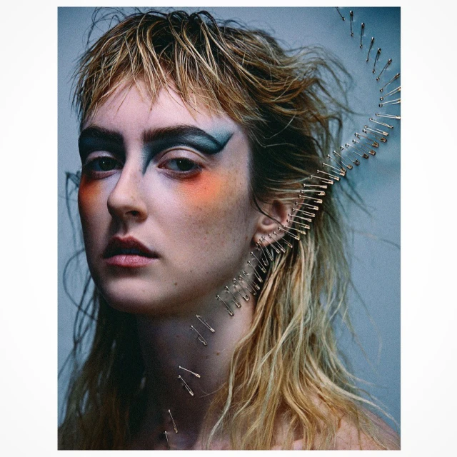 Close-up of a young woman with dramatic makeup and blonde hair, safety pins arranged around her face in a curved line.
