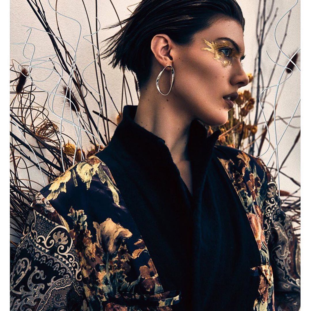 Portrait of a woman with gold eye makeup and a patterned robe, set against a backdrop of dried branches, in a beauty fashion editorial.