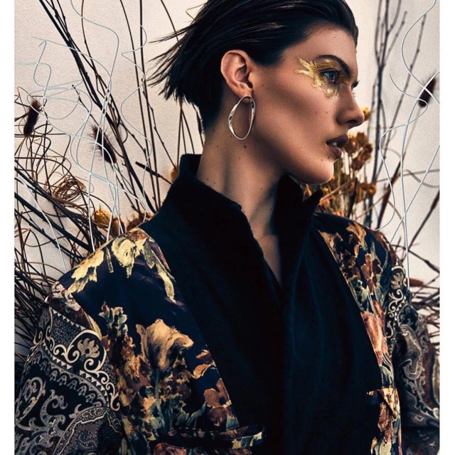 Portrait of a woman with gold eye makeup and a patterned robe, set against a backdrop of dried branches, in a beauty fashion editorial.