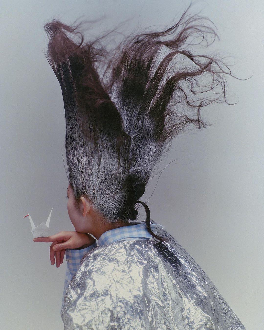 An adult woman with an elaborate updo holds a paper origami crane on her fingertip in front of a neutral background, styled for beauty or fashion.