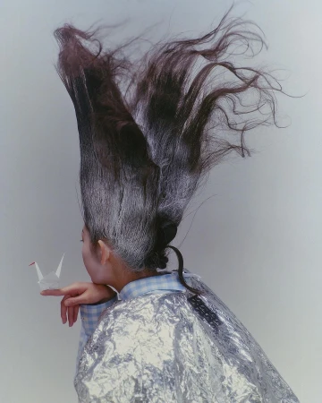 An adult woman with an elaborate updo holds a paper origami crane on her fingertip in front of a neutral background, styled for beauty or fashion.