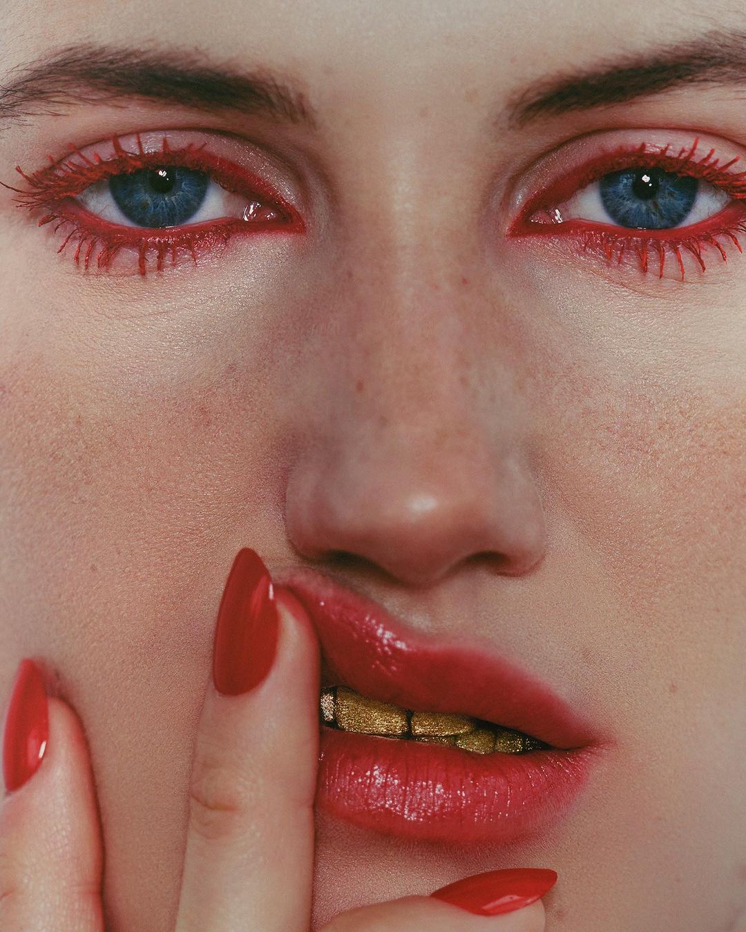 Close-up portrait of a fair-skinned adult female with blue eyes, red eye makeup, red lipstick, and gold teeth, touching her lips with red nails.