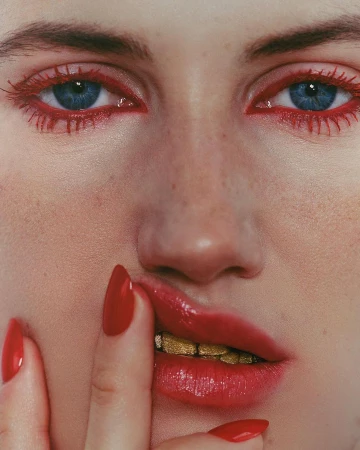 Close-up portrait of a fair-skinned adult female with blue eyes, red eye makeup, red lipstick, and gold teeth, touching her lips with red nails.