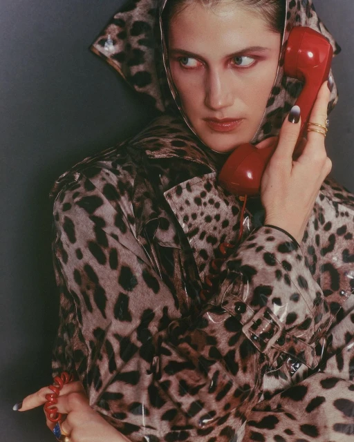 A woman in a hooded, leopard-print coat holds a red retro telephone to her ear, looking to the side against a gray backdrop.
