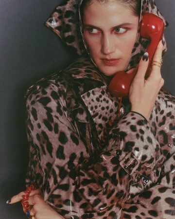 A woman in a hooded, leopard-print coat holds a red retro telephone to her ear, looking to the side against a gray backdrop.