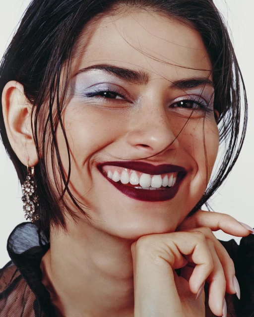 Close-up portrait of a smiling adult woman with dark hair, wearing makeup and an earring, against a bright white background.