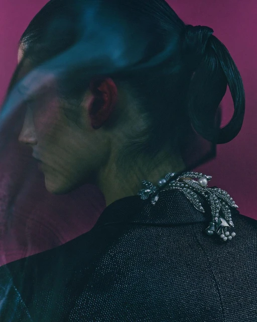 Elegant woman with ponytail wearing a textured blazer and pearl brooch against a purple background with abstract teal foreground blur.