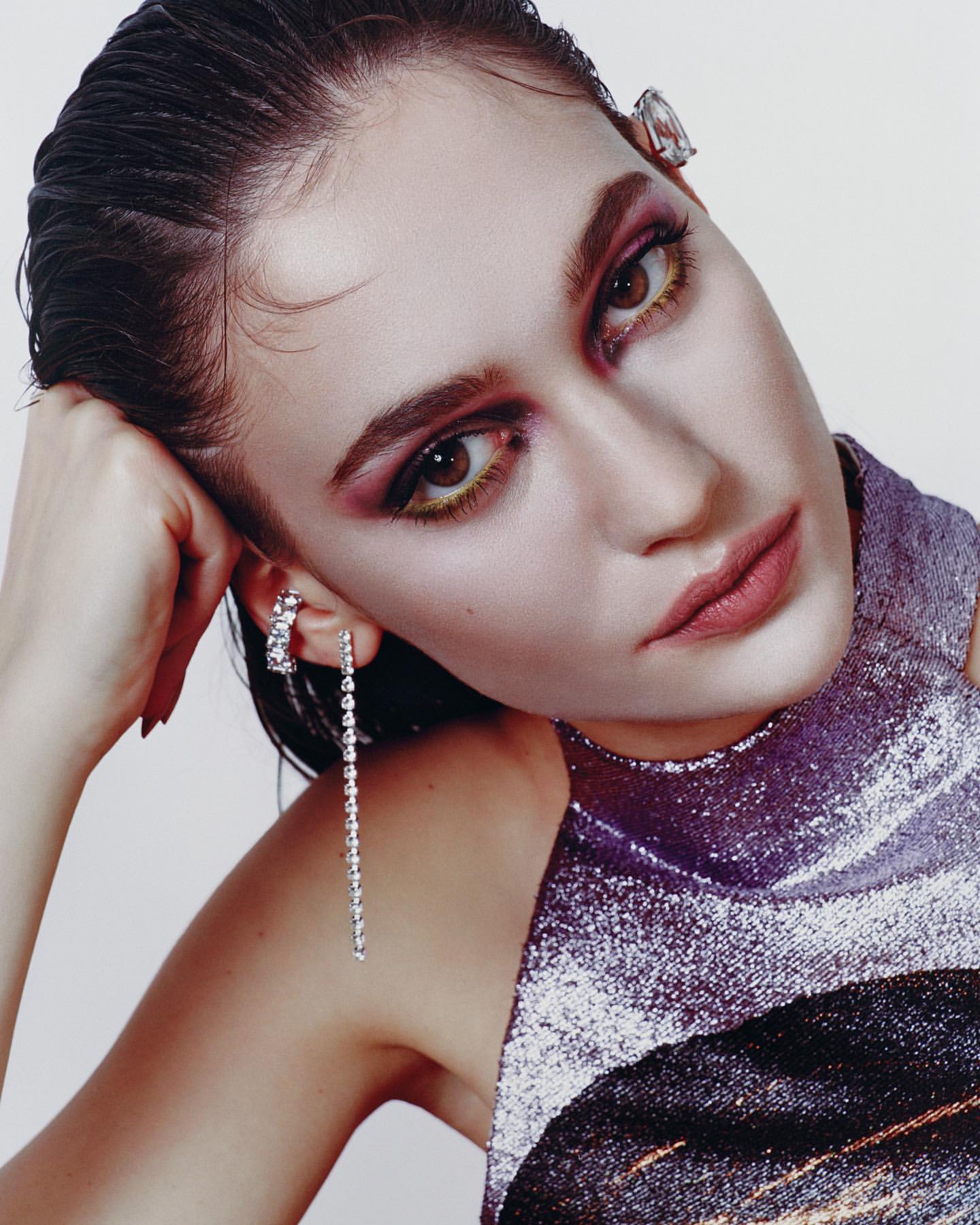 Close-up portrait of a woman with dramatic makeup and jewelry wearing a shimmering purple halter top against a plain background.