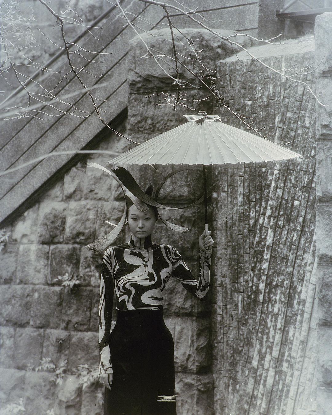 Monochrome outdoor shot of a stylish Asian woman holding umbrella, with patterned top and sleek skirt, posing against a stone structure.