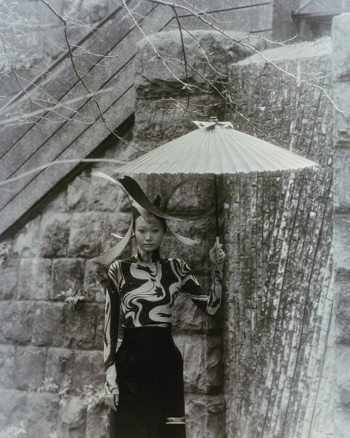 Monochrome outdoor shot of a stylish Asian woman holding umbrella, with patterned top and sleek skirt, posing against a stone structure.