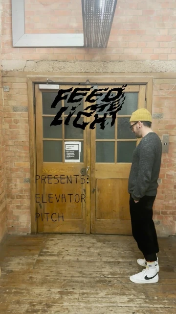 Adult male in a yellow beanie, grey sweater and black pants stands in front of doors with graphic text, next to a brick wall.