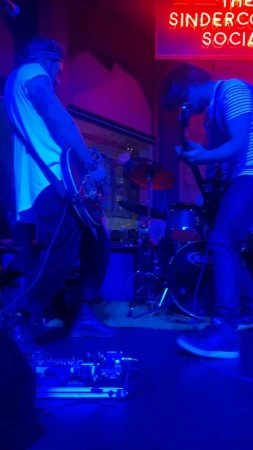 Two musicians playing electric guitars on stage with a drum set in the background under blue and pink neon lighting.
