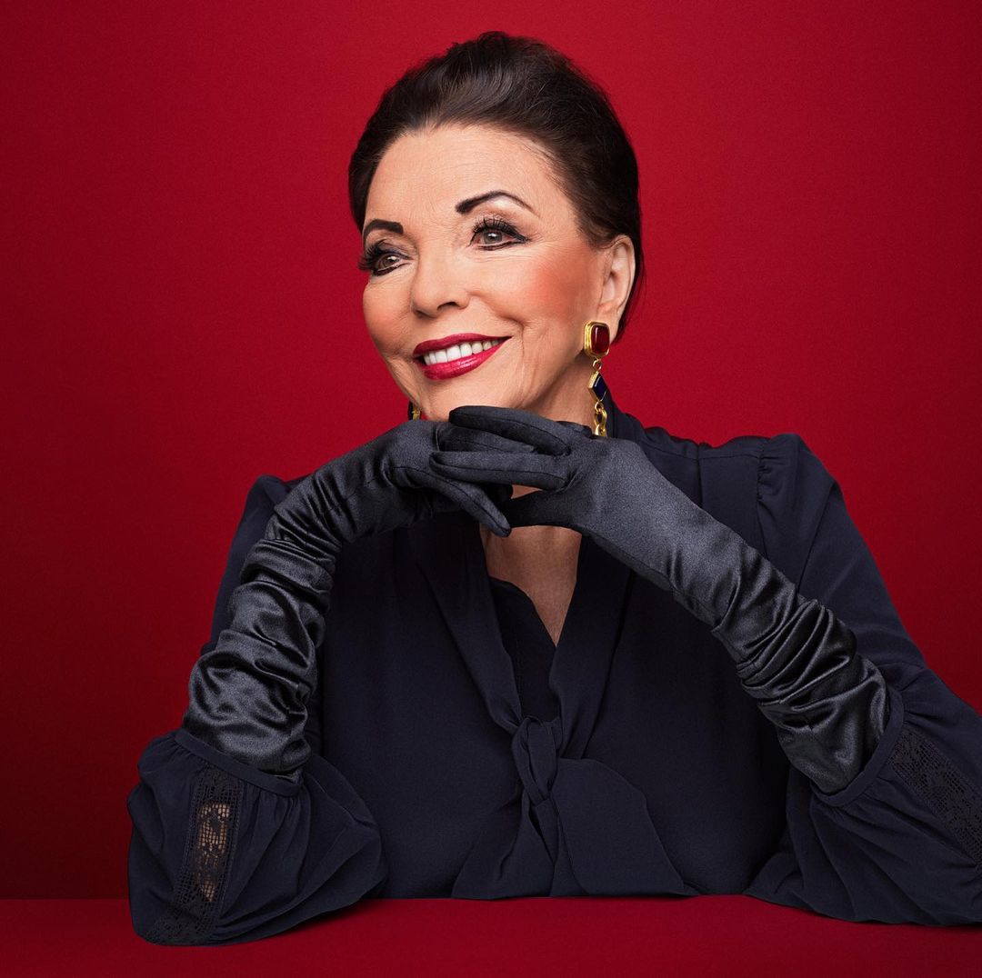 Elegant portrait of older woman with black hair and gloves against a red backdrop, featuring red lipstick, jewelry, and a sophisticated look.