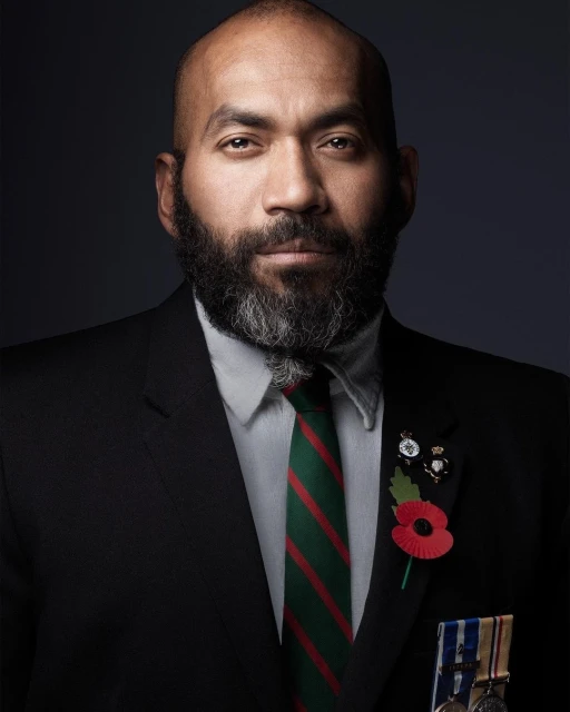 Portrait of a dignified adult male wearing a suit, white shirt, and striped tie, adorned with service pins and medals on a grey backdrop.