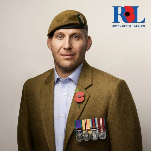 Portrait of an adult male wearing a brown tweed jacket, blue shirt, and a tan beret, featuring military medals and a poppy pin. The background is white.