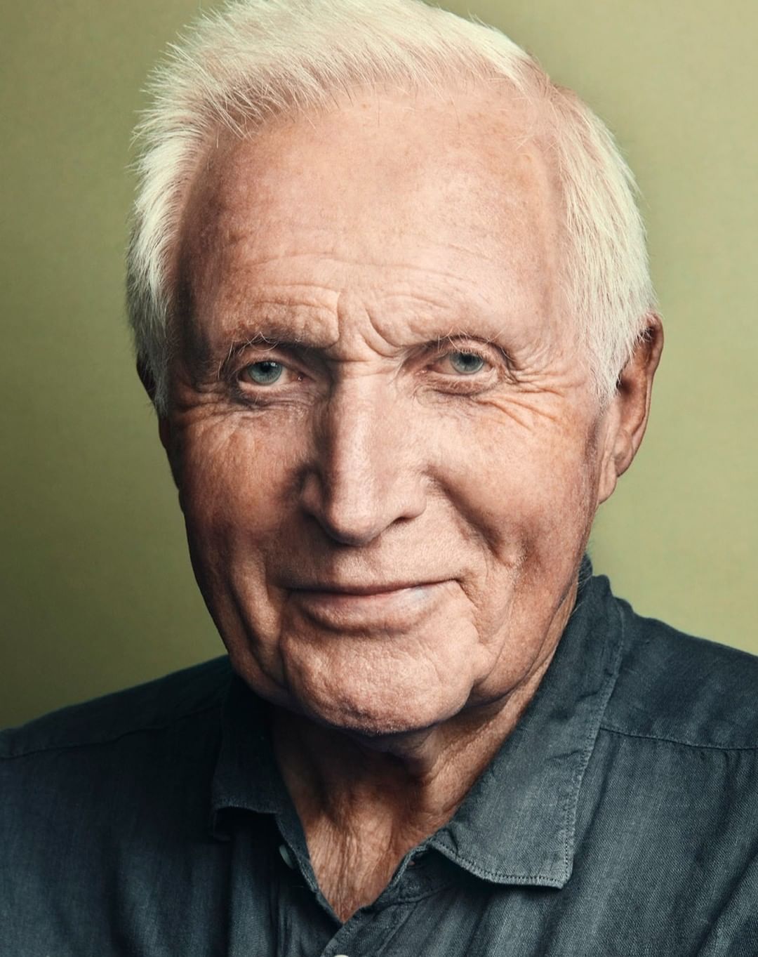 Close-up portrait of an elderly man with white hair wearing a dark grey collared shirt against a blurred background. Studio shot, head and shoulders.