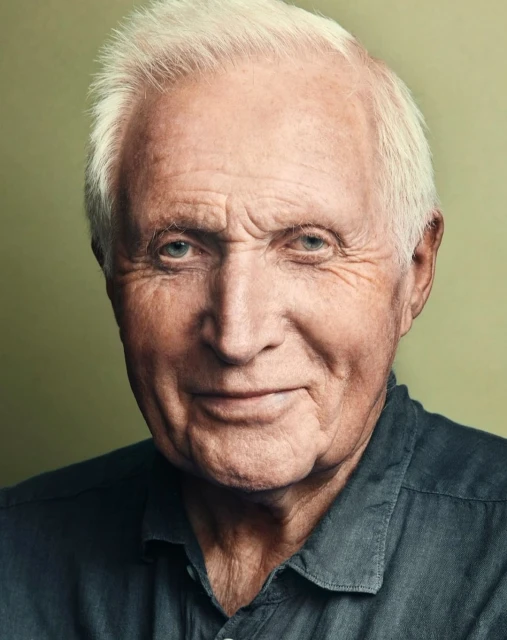 Close-up portrait of an elderly man with white hair wearing a dark grey collared shirt against a blurred background. Studio shot, head and shoulders.