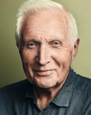 Close-up portrait of an elderly man with white hair wearing a dark grey collared shirt against a blurred background. Studio shot, head and shoulders.