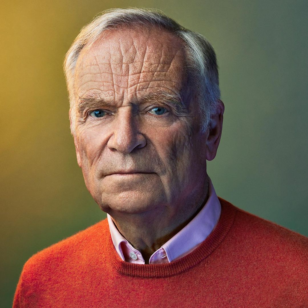 Elderly man in an orange sweater and pink collared shirt poses indoors against a blurred yellow and green background in a close-up studio shot.