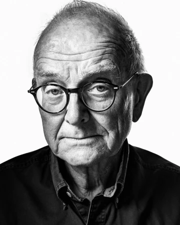 Black and white portrait of an elderly man wearing round glasses and a dark button-down shirt against a plain background.