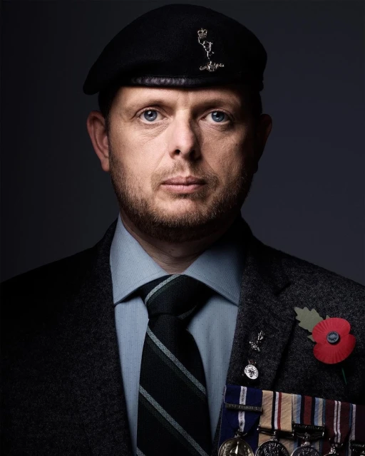 Portrait of a solemn adult male wearing a black beret, suit, tie, and military medals against a dark gray backdrop in a photo studio.