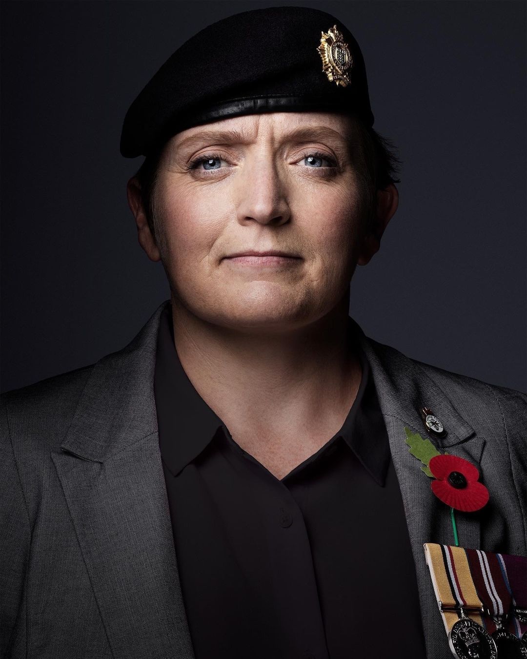 Portrait of a mature woman wearing a black beret, grey blazer, and medals against a dark background in a studio setting.