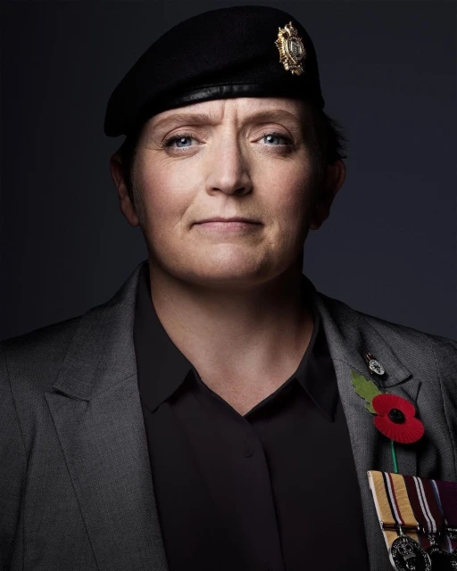 Portrait of a mature woman wearing a black beret, grey blazer, and medals against a dark background in a studio setting.