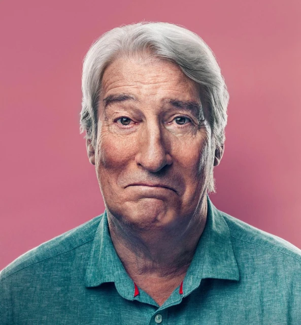 Portrait of mature adult male with grey hair, wearing a collared linen shirt, posing against a pink backdrop in a studio setting.