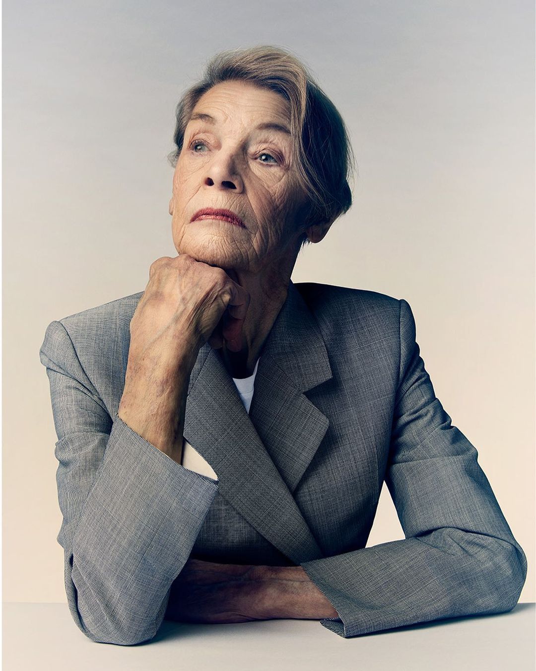 Portrait of an elderly woman wearing a grey blazer, hand to chin, looking up in a well-lit neutral indoor environment. 