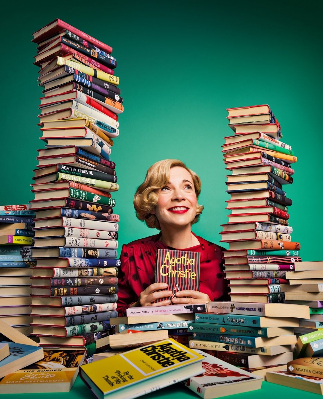 Woman with short blonde hair in a red dress smiles and holds an Agatha Christie book, surrounded by tall stacks of books against a green backdrop.