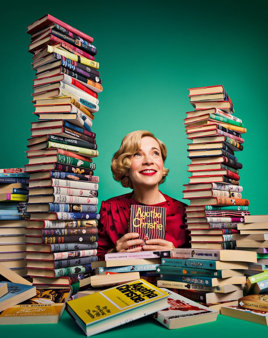 A woman with blond hair smiles, holding an Agatha Christie book, surrounded by large stacks of hardcover books against a green backdrop.